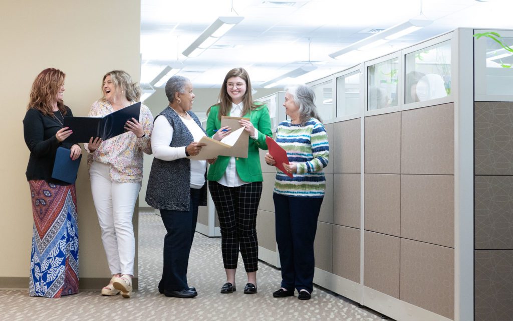 Homepage - Strate Insurance Group Team Smiling and Talking While Holding Folders with Documents as They Stand in the Hallway of their Brightly Lit Office